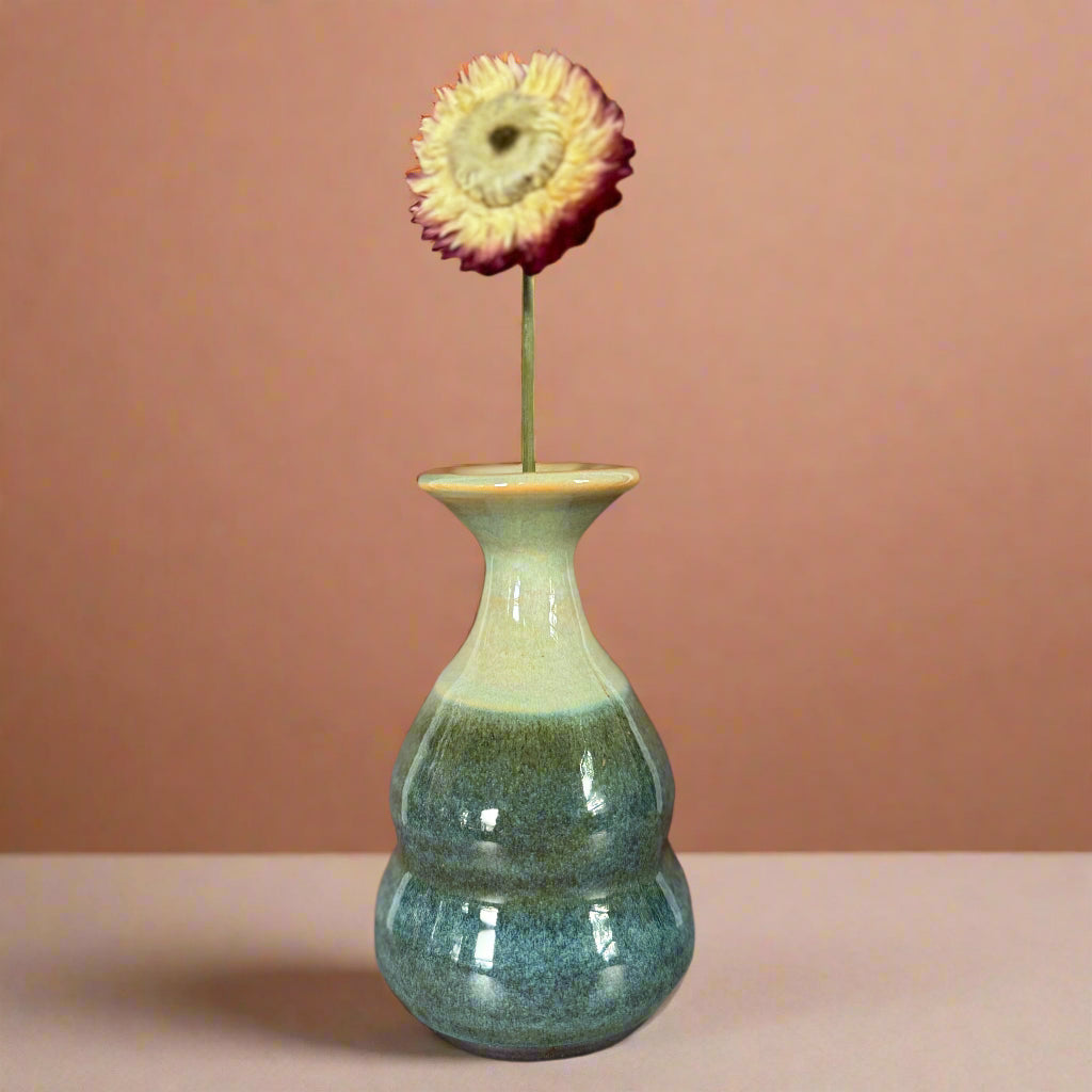 Green ceramic vase with a single flower on a wooden surface against a brown wall.