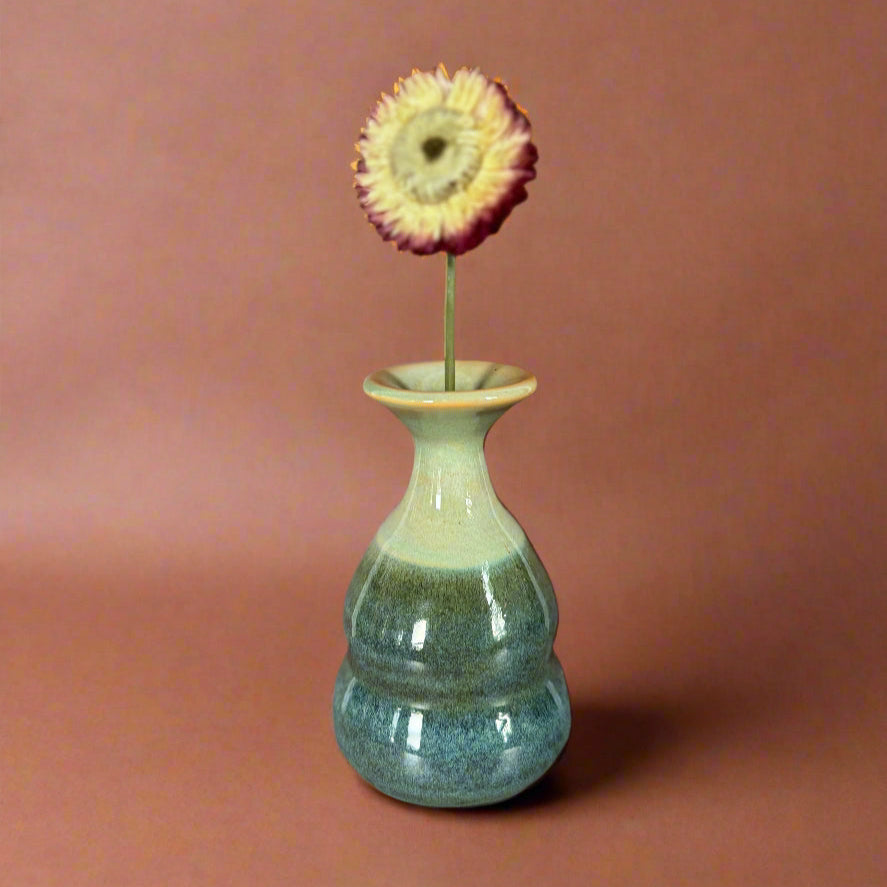 Green ceramic vase with a single flower on a wooden surface against a brown wall.