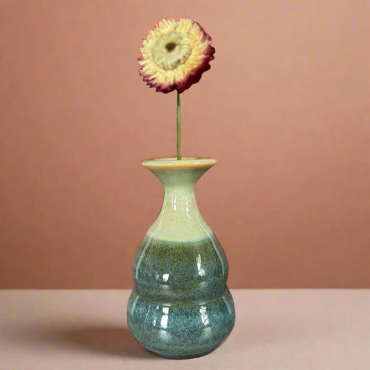Green ceramic vase with a single flower on a wooden surface against a brown wall.